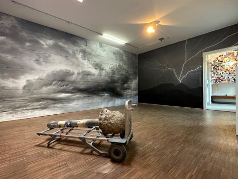 Ein Kunst Objekt von Benjamin Bergmann auf einem liegenden Transportwagen in einem Raum an dessen Wänden dunkle Regenwolken und Gewitter Blitze zu sehen sind
